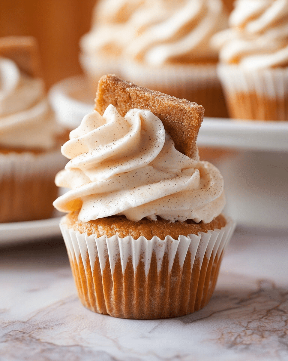 The image shows a cupcake with three main parts. The base layer is a golden-brown cupcake in a white paper liner. On top of the cupcake is a thick, soft swirl of white cream frosting with tiny specks inside it, giving it a smooth and rich texture. A small square piece of light brown cinnamon sugar toast is placed diagonally into the frosting. In the blurred background, there are more cupcakes on a white plate set on a white marbled texture. Photo taken with an iphone --ar 4:5 --v 7