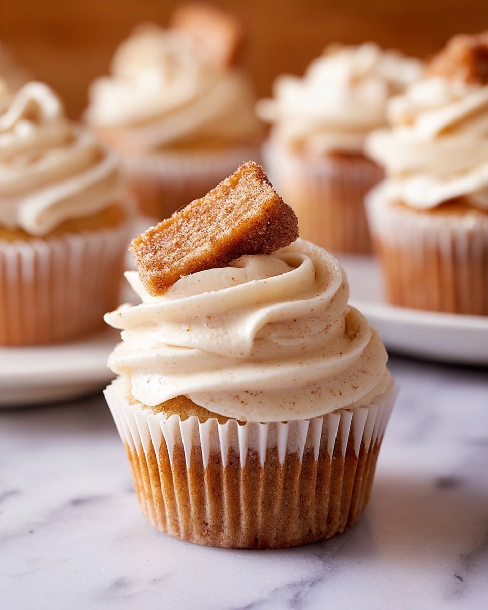 A cupcake with one visible layer of light brown cake inside a white paper liner is topped with a thick swirl of creamy off-white frosting that has tiny specks, showing texture. On top of the frosting, a small rectangular piece of spiced cake with a sugary coating is placed at an angle. In the background, a white plate holds more cupcakes with similar frosting, all set on a white marbled surface. photo taken with an iphone --ar 4:5 --v 7