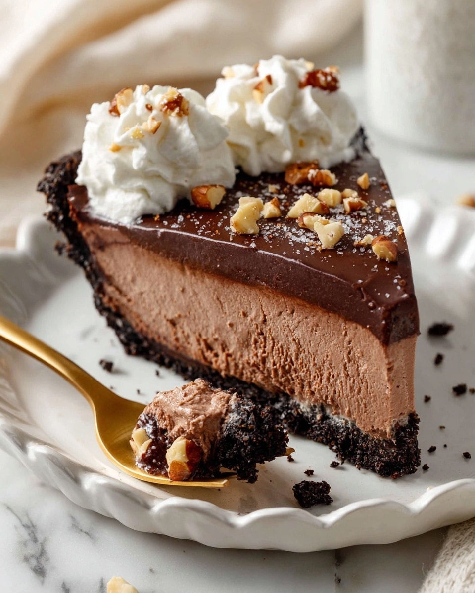 A slice of chocolate mousse pie sits on a white plate with small dark speckles and a scalloped edge, placed on a white marbled surface. The pie has three layers: a thin, crumbly dark chocolate crust at the bottom, a thick and creamy milk chocolate mousse middle layer, and a thin, glossy dark chocolate ganache layer on top. The ganache is sprinkled with small chunks of toasted nuts and flakes of sea salt. Two swirls of soft white whipped cream sit at the upper back edge of the slice. In front of the pie, a gold fork holds a bite-sized piece of the mousse with bits of crust and nuts. Photo taken with an iphone --ar 4:5 --v 7