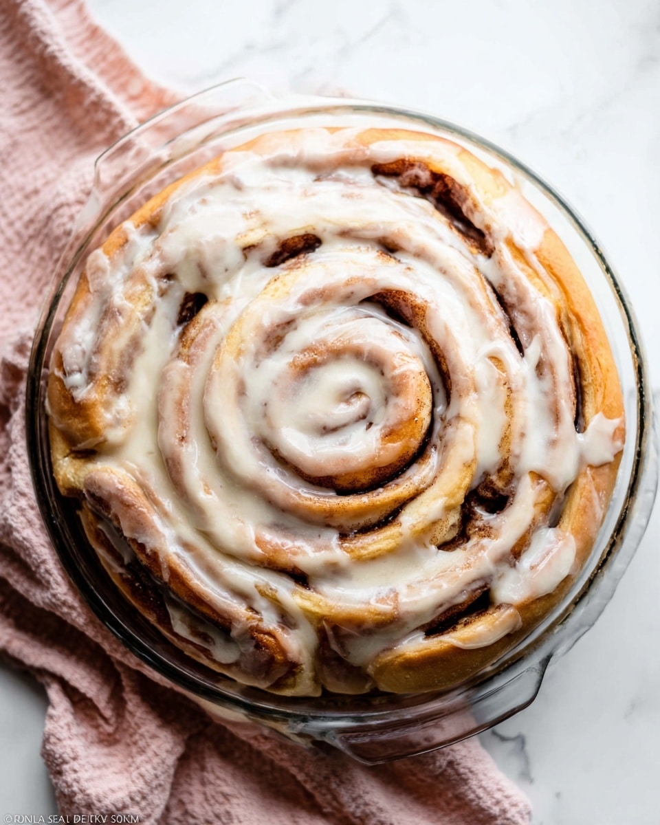 A large cinnamon roll is shown from above, shaped in a tight spiral with multiple layers of fluffy, golden-brown dough and dark cinnamon filling visible between each layer. The roll is generously covered with a creamy white icing that drips slightly down the sides, giving it a glossy and smooth texture. The cinnamon roll sits inside a clear glass pie dish resting on a soft light pink and white cloth, all placed on a white marbled surface. The icing looks thick and soft, blending nicely with the warm tones of the cinnamon and dough. photo taken with an iphone --ar 4:5 --v 7