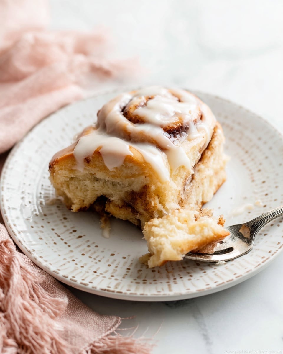 A piece of cinnamon roll sits on a white plate with small dot patterns around the edge, placed on a white marbled surface. The cinnamon roll has four visible layers of soft, light golden dough spiraled with a dark cinnamon filling. The top is covered in a smooth, creamy white icing that drips slightly down the sides. Next to the cinnamon roll, a silver fork holds a small piece of the roll with some icing. A light pink cloth with fringes is partially visible at the edge of the image. photo taken with an iphone --ar 4:5 --v 7