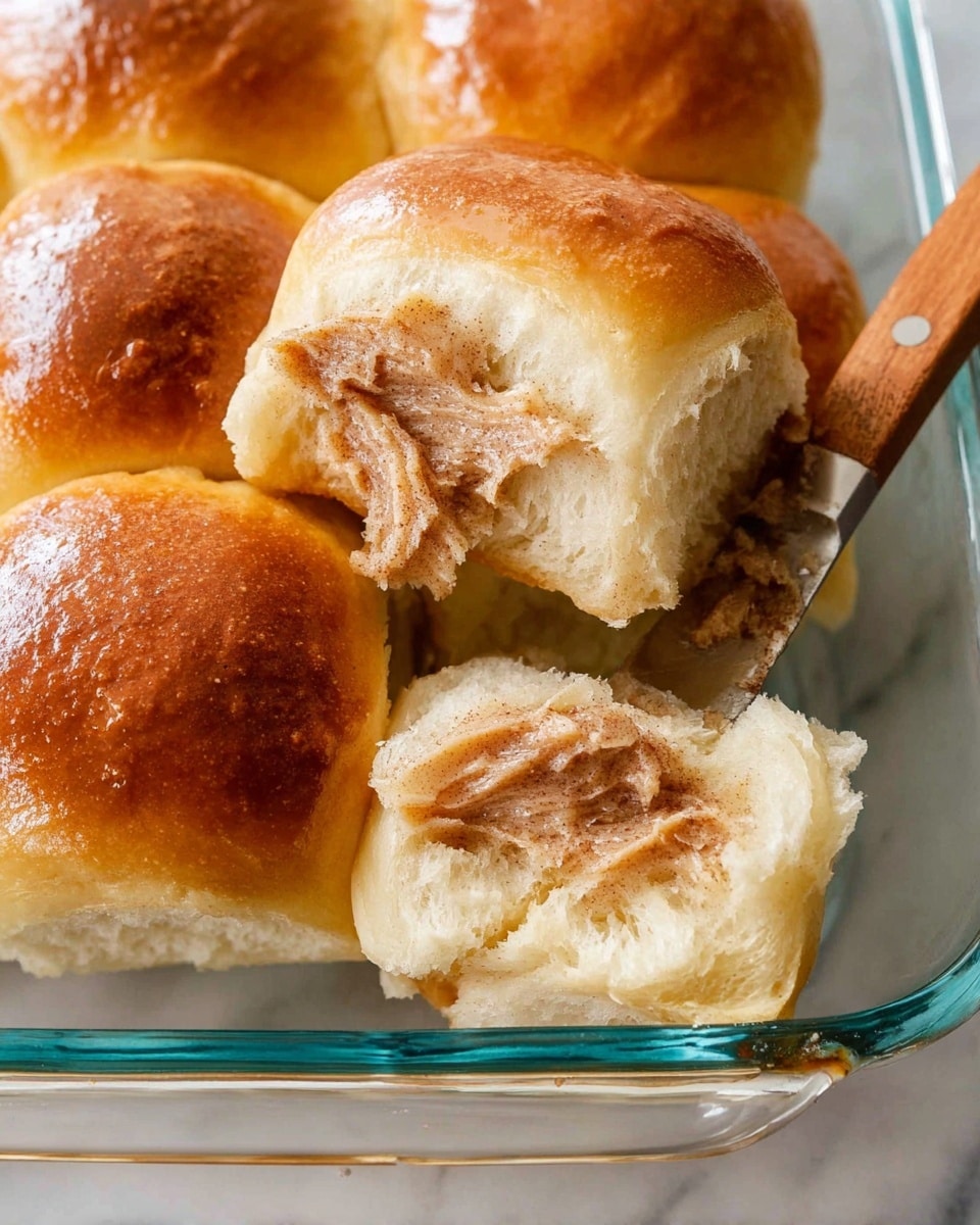 A close-up view of a group of five soft, golden-brown dinner rolls placed closely together inside a clear glass baking dish, showing the fluffy texture of the bread. Two rolls in the front have been pulled apart slightly, revealing the soft, airy inside spread with a creamy light brown cinnamon butter that has a smooth texture with darker cinnamon specks. A butter knife with a wooden handle, coated lightly with the same cinnamon butter, rests on the edge of the glass dish. The dish is set on a white marbled surface photo taken with an iphone --ar 4:5 --v 7