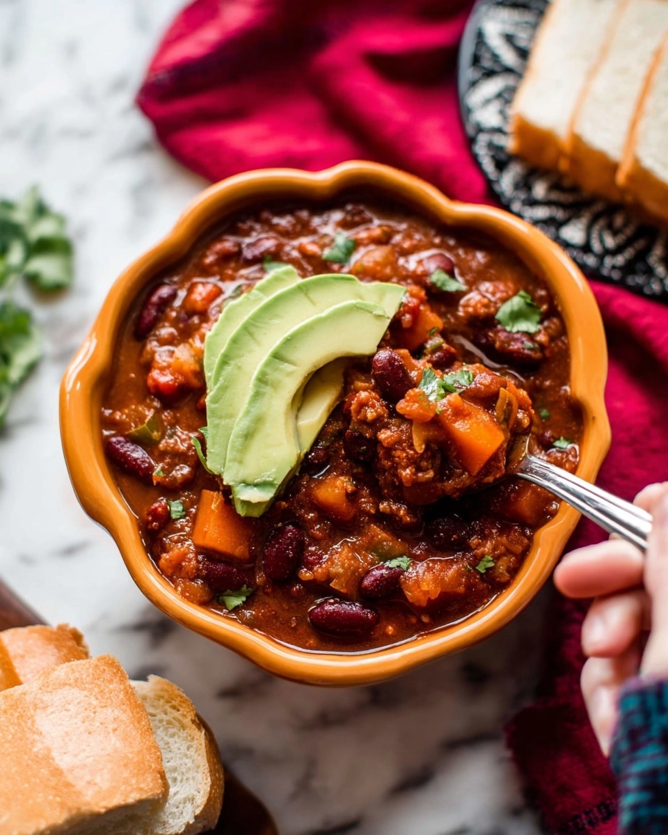 The image shows a bowl of thick chili in an orange scalloped bowl sitting on a white marbled surface. The chili has layers of rich brown and red chunky sauce mixed with dark red kidney beans, small diced orange and red veggies, and some green cilantro leaves peeking through. On top are two slices of smooth, light green avocado. A woman's hand holds a spoon scooping the chili from the right side. In the background, there is white sliced bread on a dark patterned plate and a red cloth. photo taken with an iphone --ar 4:5 --v 7