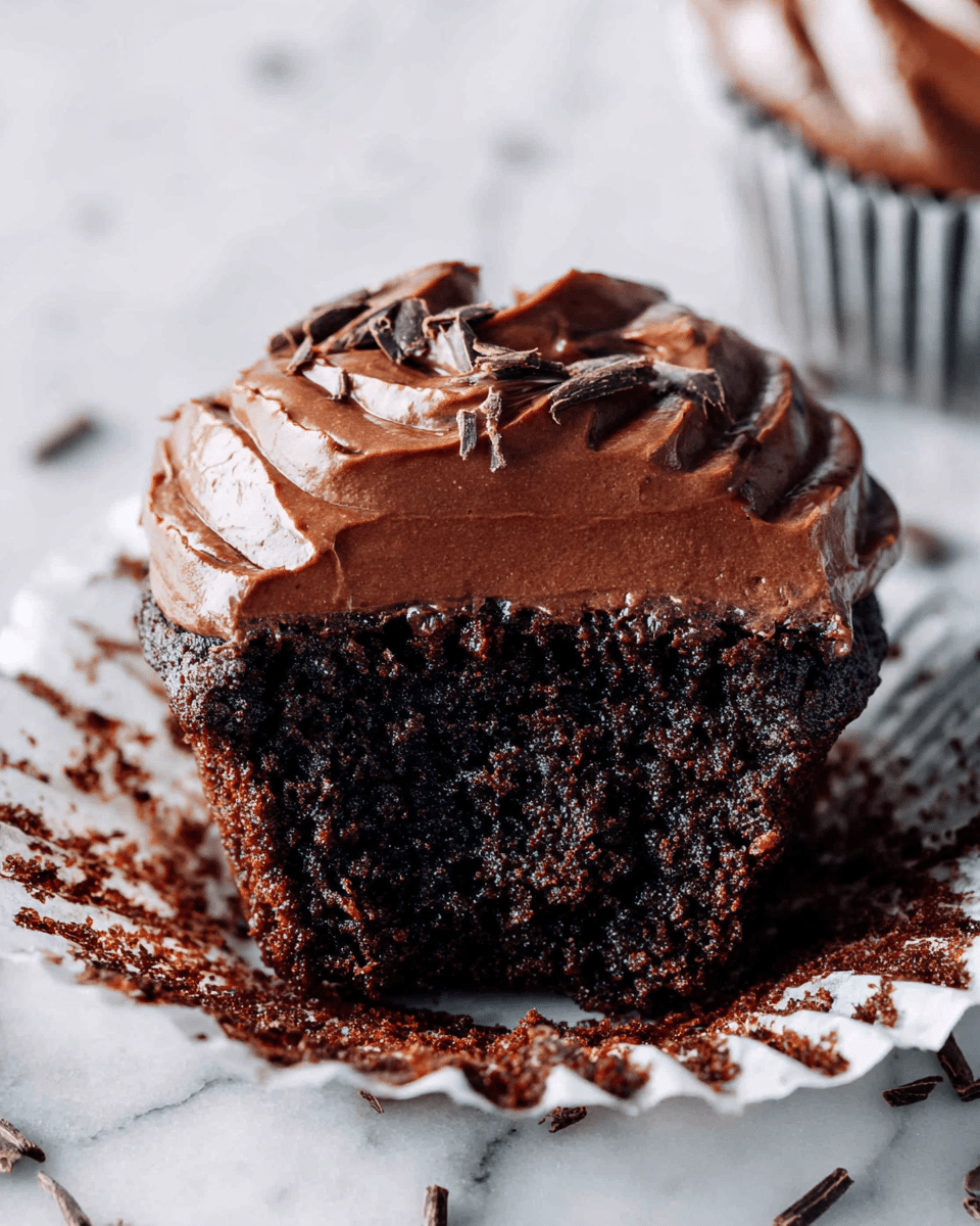 A rich chocolate cupcake with two layers: the bottom is a dark, moist chocolate cake with a rough texture, and the top layer is smooth, creamy chocolate frosting that has a glossy shine and ridged pattern. The cupcake sits in a white paper liner stained with melted chocolate, and small chocolate shavings are scattered lightly on the frosting and around the edges. The background is a white marbled texture, adding a clean and bright contrast to the dark chocolate colors. Photo taken with an iphone --ar 4:5 --v 7