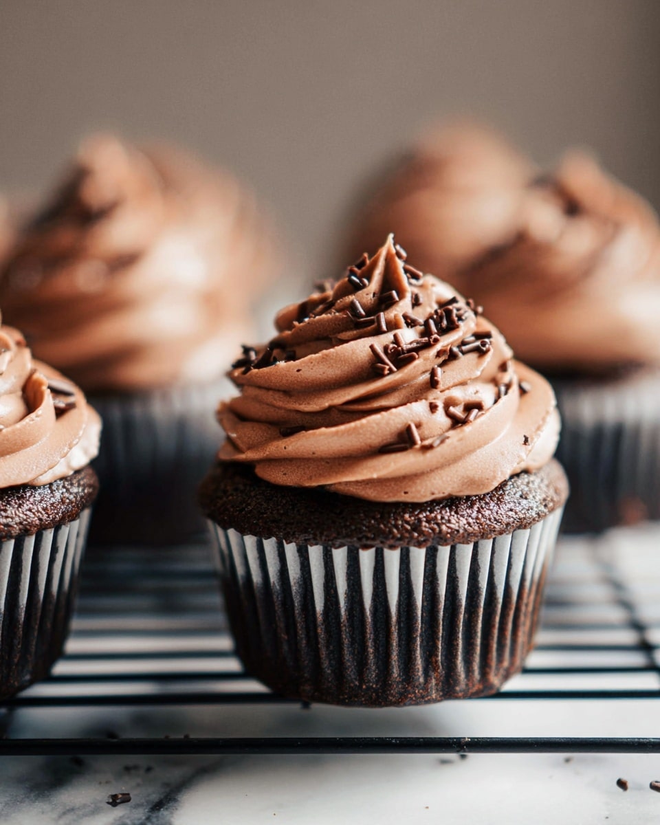 A close-up of multiple chocolate cupcakes placed on a black wire rack over a white marbled surface, each cupcake topped with three thick swirls of smooth, creamy milk chocolate frosting. The frosting is sprinkled with small chocolate curls that add texture and decoration, creating a rich and inviting look. The cupcake base is dark chocolate-colored, with a slightly textured surface and a black and white striped paper liner. The image focuses on one cupcake sharply, while the others are softly blurred in the background, with natural light casting gentle shadows. photo taken with an iphone --ar 4:5 --v 7