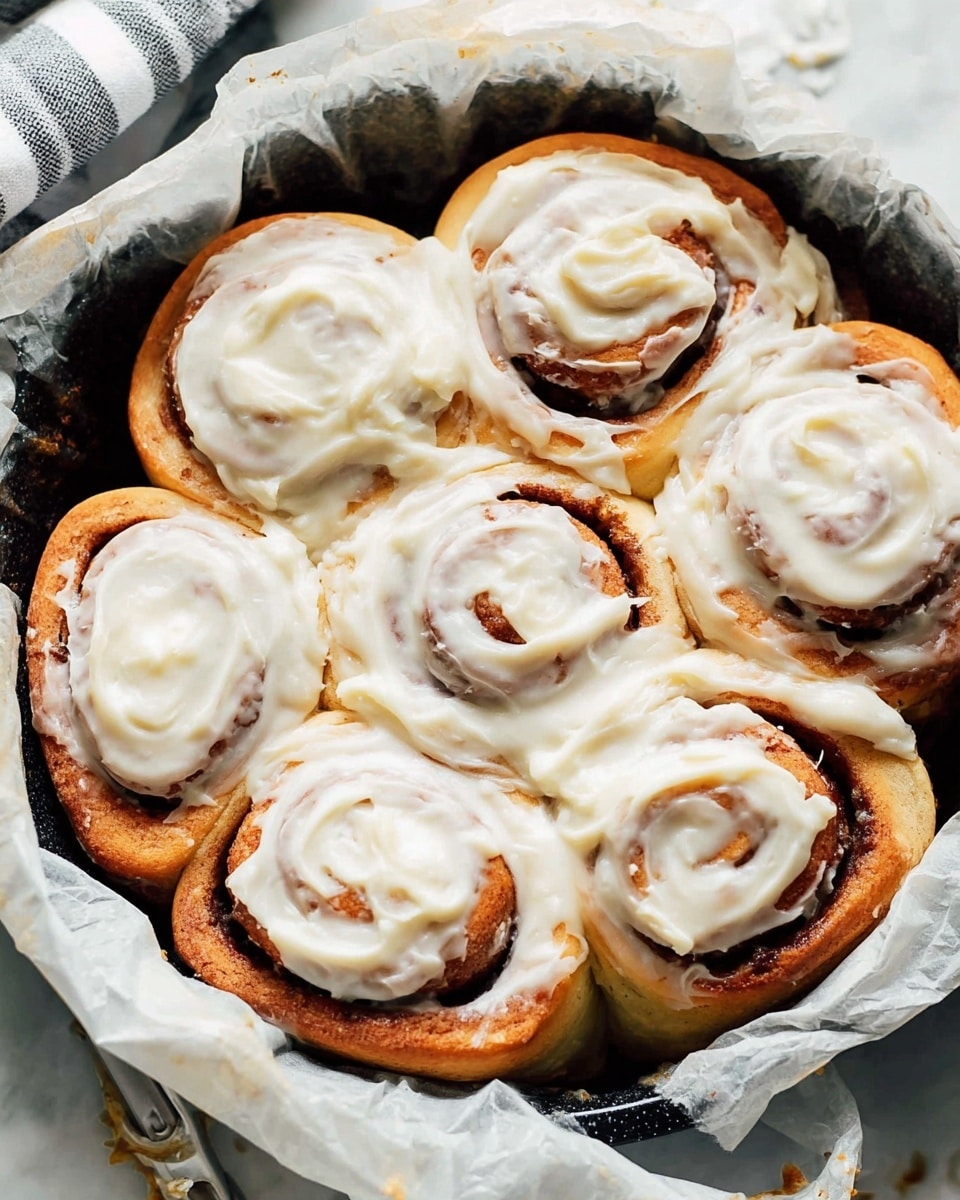 A close-up view of six cinnamon rolls tightly placed in a round black pan lined with crumpled white parchment paper, each roll showing a golden-brown spiral layer of dough with dark brown cinnamon filling inside; the rolls are generously covered with a thick layer of creamy white frosting that has a smooth and slightly glossy texture, having tiny specks of vanilla; the swirls of the cinnamon rolls peek through the frosting in places, giving a contrast between the warm brown tones and the bright white icing; the pan rests on a white marbled surface with a hint of a striped cloth in the top corner photo taken with an iphone --ar 4:5 --v 7