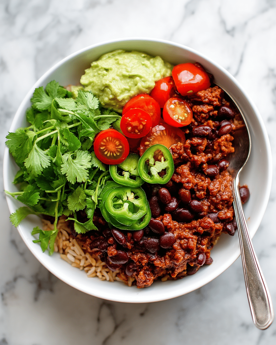 A white bowl filled with a base layer of light brown rice mixed with black beans, topped with a thick layer of dark red chili made of ground meat and beans. On top of the chili, there is a mix of fresh bright green cilantro leaves, bright red cherry tomatoes cut in halves, and several slices of green jalapeño peppers. To the side, a dollop of light green guacamole with a smooth texture is visible. A silver spoon is partially inserted into the rice on the right side of the bowl. The bowl sits on a white marbled texture surface. photo taken with an iphone --ar 4:5 --v 7