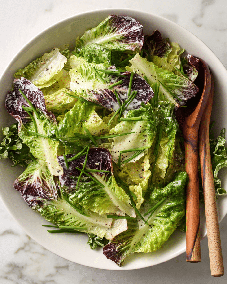 A bowl filled with layered salad leaves including light green and darker green shades, some leaves have a hint of purple. The leaves are lightly coated with a creamy dressing, speckled with ground black pepper. On top, fresh green chive pieces are scattered evenly. A pair of salad servers with wooden handles rests on the edge of the white bowl, which sits on a white marbled surface. photo taken with an iphone --ar 4:5 --v 7