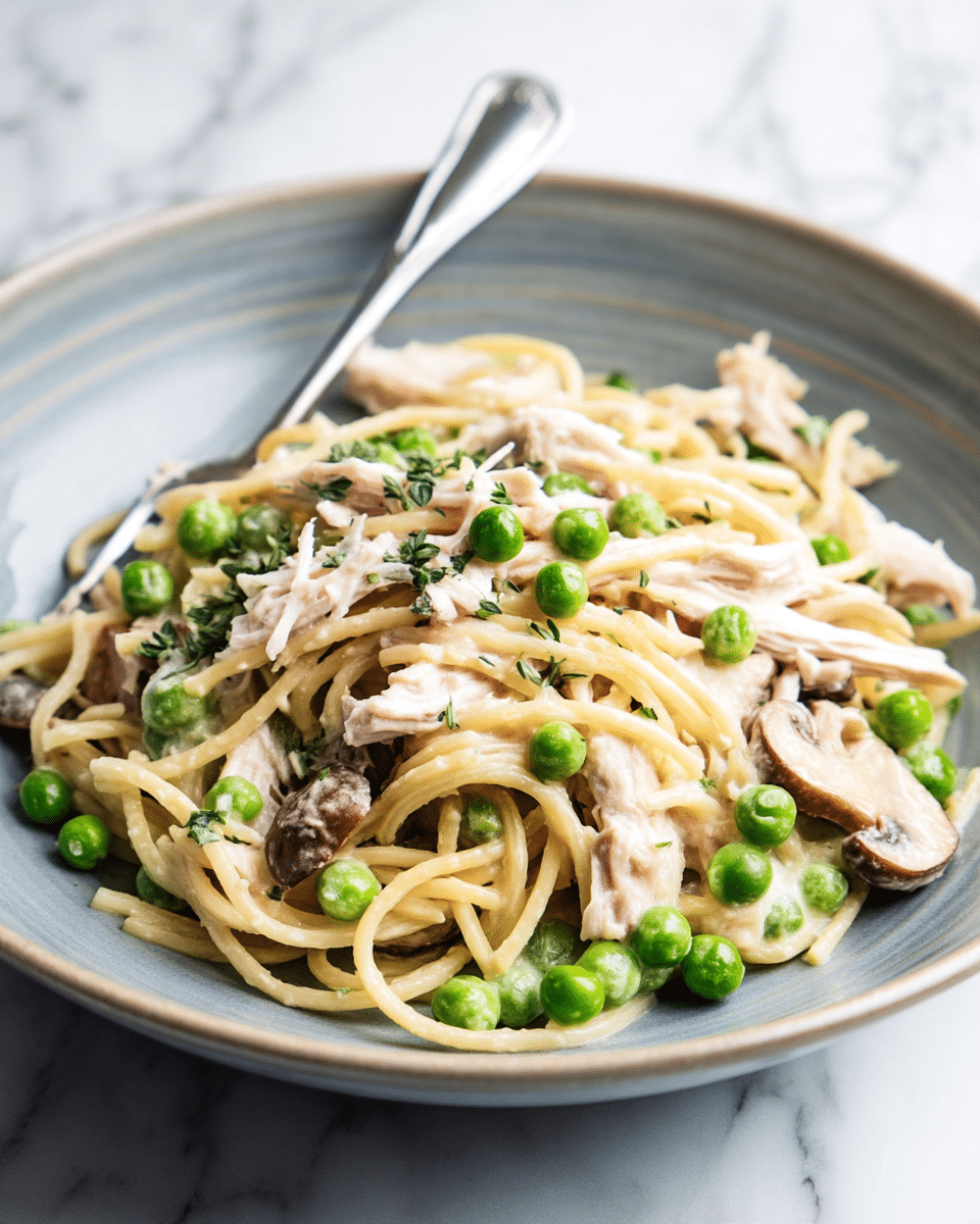 A close-up of a bowl of creamy pasta with three main visible layers: at the base, light beige spaghetti noodles tangled and coated in a creamy white sauce; mixed throughout are bright green peas and light brown sliced mushrooms with a soft texture, along with shredded white chicken pieces scattered evenly on top. The bowl is a wide, shallow ceramic bowl in pale grayish blue with a matte finish, set on a white marbled textured surface. A silver fork is partly visible inserting into the pasta from the left side. Photo taken with an iphone --ar 4:5 --v 7