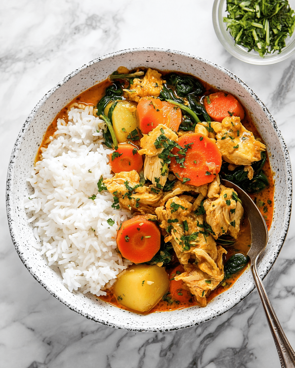 A white speckled bowl holds a dish with two main layers: one side filled with fluffy white rice, and the other side covered in a rich orange curry with tender pieces of light-colored chicken, bright orange carrot slices, yellow potato chunks, and dark green spinach leaves. The curry sauce looks creamy and is garnished with small fresh green herb pieces. A silver spoon rests on the right side of the bowl, partially scooping some chicken and spinach. In the background, a small white bowl with chopped green herbs can be seen on a white marbled surface. photo taken with an iphone --ar 4:5 --v 7