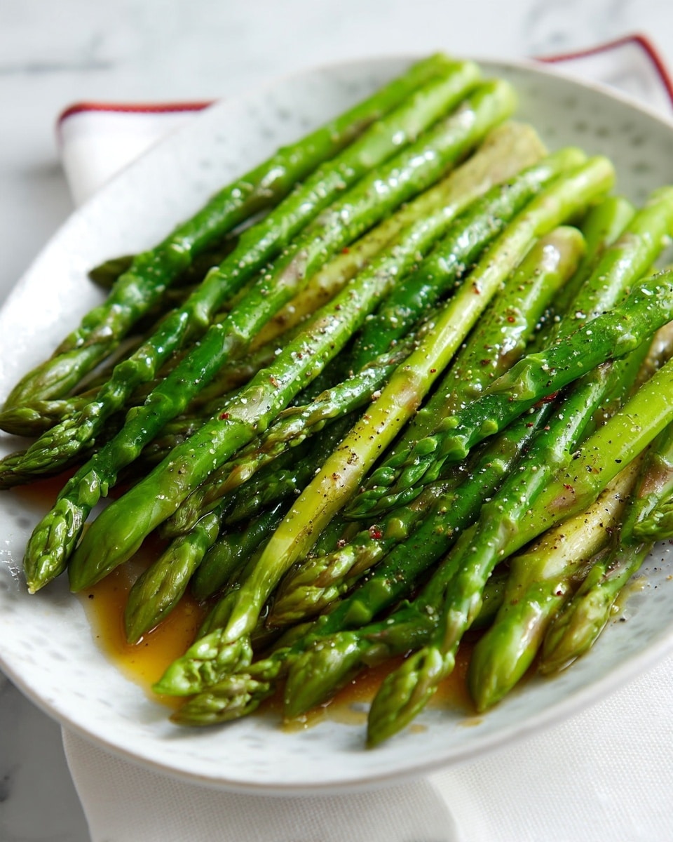 A white plate filled with about two dozen cooked asparagus stalks arranged closely and overlapping each other. The asparagus stalks are bright green with slightly darker tips and lighter, almost white lower stems. Light oil and seasoning with visible specks of black pepper and coarse salt coat the stalks, giving them a shiny, slightly wet texture. The plate rests on a white cloth napkin with a narrow red border, all set on a white marbled surface. photo taken with an iphone --ar 4:5 --v 7