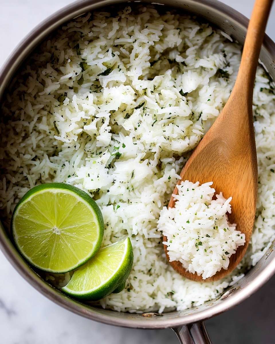 Inside a shiny stainless steel instant pot, there is a layer of white rice mixed with small bits of green herbs, giving it a light speckled texture. On the left side of the rice, three wedges of bright green lime rest close together. Sticking into the rice from the right side is a light brown wooden spoon, its smooth surface visible and blending with the texture of the rice. The pot sits on a white marbled surface, with a small white bowl below and slightly to the right holding a few lime wedges. To the bottom right, green fresh cilantro leaves peek into the frame, and a dark blue textured cloth lies crumpled to the lower left of the pot. The top right corner has black and gray text that reads