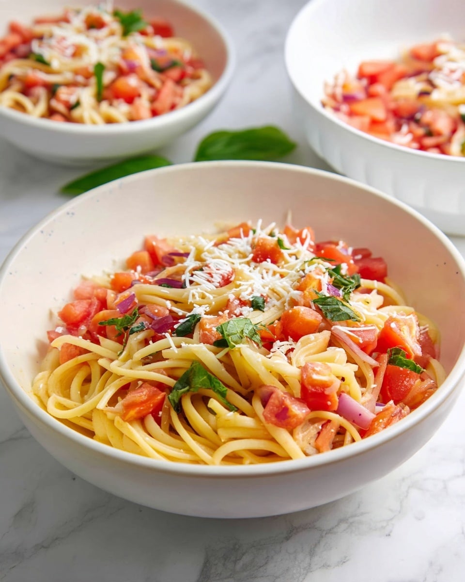 A close-up view of a white bowl filled with spaghetti pasta tossed with small, chopped red tomatoes and green basil leaves scattered throughout. The pasta strands are light yellow and appear soft and cooked. The top layer is sprinkled with fine white grated cheese, adding a light texture contrast. In the background, parts of two more white bowls filled with similar pasta mixes are visible on a white marbled surface. The image highlights fresh, colorful ingredients with a simple, clean presentation. photo taken with an iphone --ar 4:5 --v 7