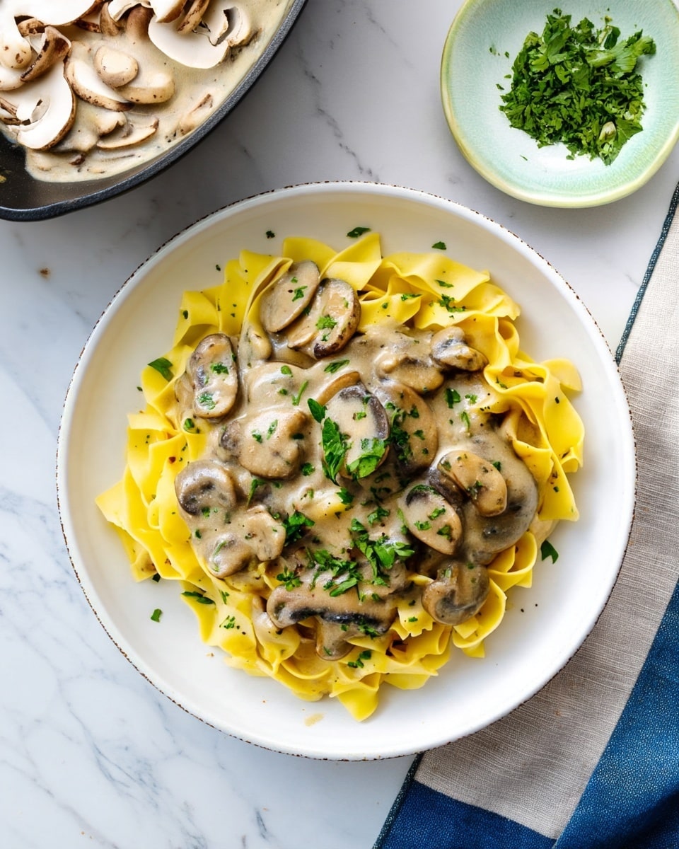A white bowl holds a serving of wide, yellow pasta ribbons as the bottom layer, topped with a creamy beige mushroom sauce thickly covering the pasta. Sliced mushrooms, light brown with darker edges, are mixed into the sauce, and fresh green chopped herbs are sprinkled over the top, adding a touch of color. Around the bowl, there is a white marbled surface holding a skillet with more mushroom sauce visible in the top left corner, a small white plate with chopped herbs in the top right, and parts of another plate with raw mushroom slices and a beige cloth with a blue edge at the bottom right. Photo taken with an iphone --ar 4:5 --v 7