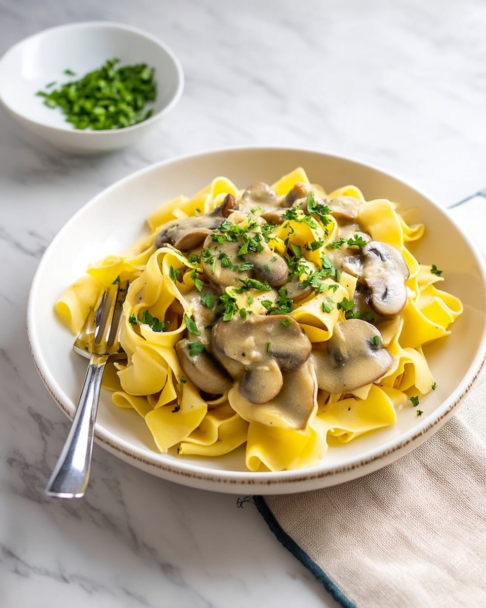 A round beige plate holds a serving of wide, yellow pasta ribbons arranged in a loose pile. On top, there is a thick, creamy mushroom sauce with visible sliced mushrooms and a slightly glossy texture. Each mushroom slice is coated in the light brown sauce, and the dish is sprinkled with small bits of chopped green herbs. To the top right of the plate, a small bowl filled with more chopped green herbs rests on a white marbled surface. The overall presentation is clean and simple, with soft natural lighting. Photo taken with an iphone --ar 4:5 --v 7