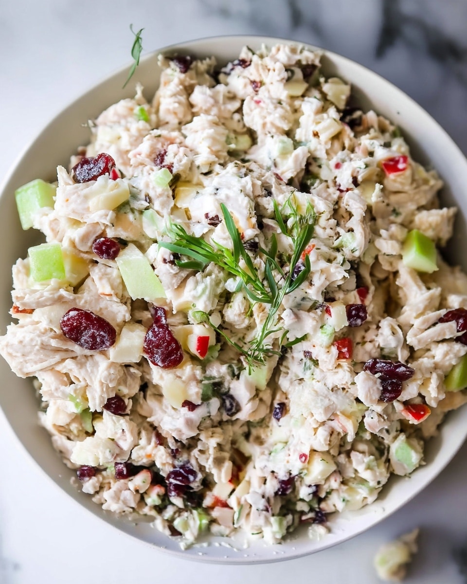 A close-up image of a creamy chicken salad served in a deep white bowl, placed on a white marbled surface. The salad has multiple small layers of white shredded chicken mixed with chunky pieces of light green celery, small cubes of red and green apple, dark reddish-brown dried cranberries, and some tiny light beige nuts, all coated in a creamy white dressing. In the center, a small sprig of fresh green herbs adds a pop of color and freshness to the dish. photo taken with an iphone --ar 4:5 --v 7