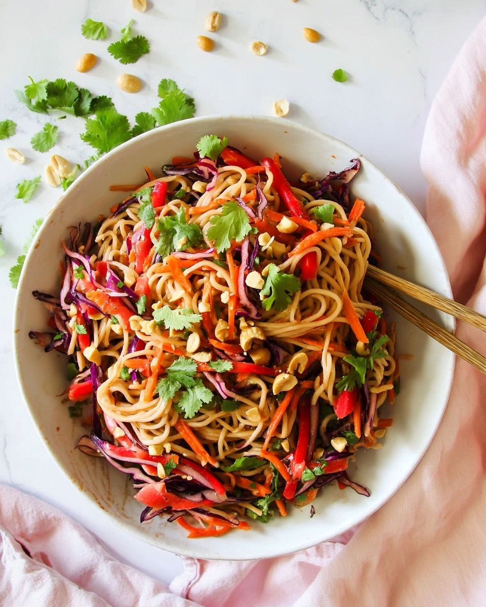 A large white bowl filled with a colorful noodle salad featuring three main layers: light brown noodles forming the base, topped with thin strips of bright orange carrots, deep purple red cabbage, and red bell peppers, all mixed evenly. The salad is scattered with fresh green cilantro leaves and small pieces of light tan peanuts, adding texture. The bowl sits on a clean white marbled surface with a few loose cilantro leaves and peanuts around it. At the right side of the bowl, a pair of wooden chopsticks rests partially inside the salad. A soft pink cloth is placed below the bowl. Photo taken with an iphone --ar 4:5 --v 7