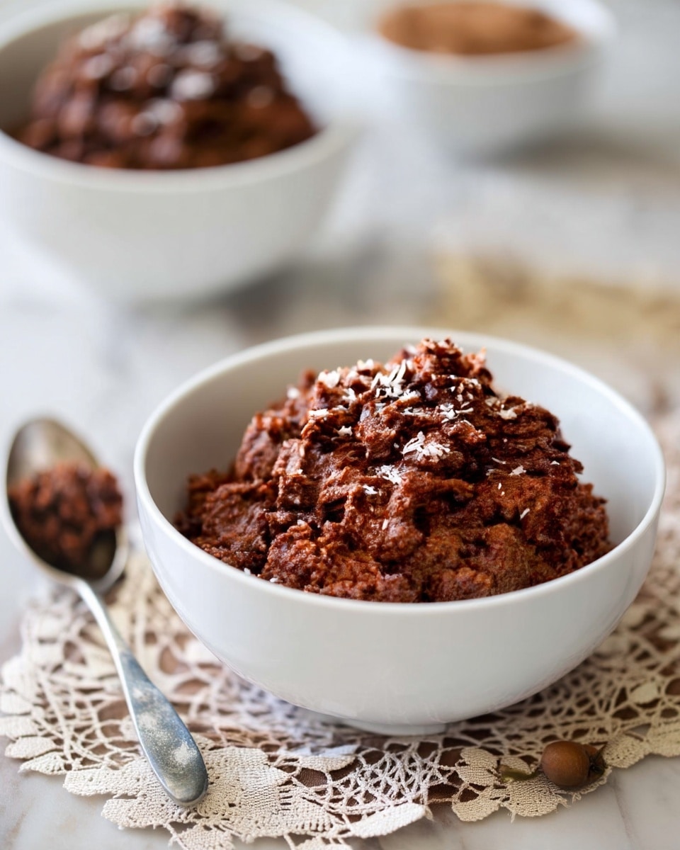 A white bowl filled with a mound of dark brown crumbled mixture, topped lightly with white flakes, sitting on a beige lace cloth. In the blurred background, there are two more white bowls with similar brown contents, placed on a white marbled surface. A metal spoon lies next to the bowl in the foreground. photo taken with an iphone --ar 4:5 --v 7